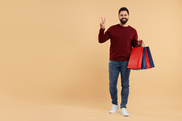 Sticker - Smiling man with many paper shopping bags showing peace sign on beige background. Space for text