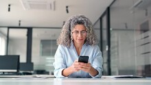 Happy middle aged business woman holding mobile cell phone using cellphone in office. Smiling mature older professional lady business owner entrepreneur using smartphone working sitting at desk.