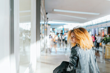 Canvas Print - A woman with curly hair is walking through a mall. She is wearing a black jacket and she is looking at windows. The mall is crowded with people, and there are several potted plants and a bench in the 