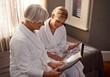 © MV/peopleimages.com - Grandmother, woman and hospitality at home, hotel and happy for bonding and looking at photo album memories. Senior lady, daughter and moment for self care, robe and bedroom for comfort and spa day