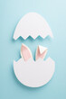 Delightful Easter surprise concept. Overhead vertical shot of amusing bunny ears emerging from a broken egg on a soft blue canvas, leaving space for festive messages or ads