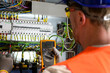 © juraj - electrician in blue helmet, yellow glasses and orange vest measures electric current with digital multimeter in distribution box-blurred background