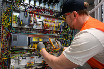 an electrician in a cap, yellow glasses and an orange vest measures electric current with a digital multimeter on a distribution box