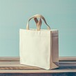 This image depicts a simple, cream-colored tote bag with a sturdy, woven pair of handles, standing upright on a horizontal wooden surface. The bag is empty and appears to be made of a canvas-like fabr