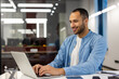 © Tetiana - Smiling young Muslim man businessman, designer, freelancer, programmer in blue shirt working in office with laptop, sitting at office table and typing on keyboard