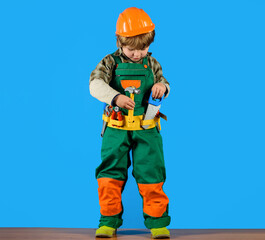 Wall Mural - Little builder child playing with toy hammer and saw. Child game. Small boy in overalls and hard hat with builder toy tools set. Cute kid in protective helmet with toolbelt of toy tools for repair.