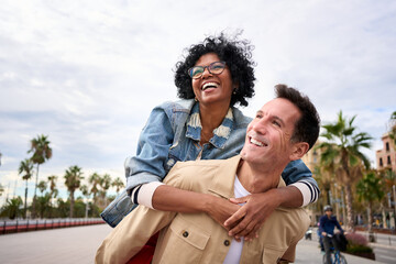 Wall Mural - Mature multiracial joyful tourist couple piggybacking standing outdoor. Cheerful black woman and Caucasian man smiling walking on city street enjoying happy vacation. Copy space, background palm trees