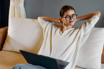 Wall Mural - Smiling woman using laptop on the cozy couch in her modern home office The attractive freelancer is enjoying her online work, surrounded by an inviting indoor atmosphere With her glasses on, she types