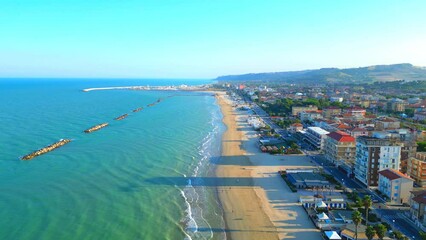 Wall Mural - Splendid aerial pullback shot from Porto San Giorgio (Fermo) of the Adriatic Sea with dark lime reflections, foamy waves reaching the shore, breakwaters, dock, buildings, roads with cars, hillscapes