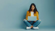 © Studio Nova - A young woman in a yellow sweater and jeans sits on the floor with her legs crossed, engrossed in her laptop.
