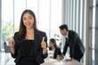 © Bavorndej - Asian businesswoman gives a thumbs up to the camera with an attractive smile. She stands with team at the office.