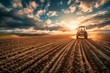 A tractor plows the field, creating deep furrows in the soil against a dramatic sunset backdrop, symbolizing agriculture and hard work