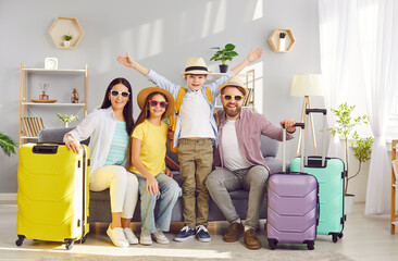 Happy cheerful parents with children daughter and son sitting on sofa with packed suitcases and looking at camera ready to fly to tropical resort together. Family vacation and holiday trip.