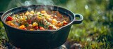 Fototapeta Uliczki - A cast iron pot filled with a hearty soup made of chicken, tomatoes, onions, and peppers is placed on top of a lush green grassy field under the open sky.