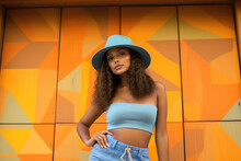Elegant fashion model in a turquoise hat and crop top poses against an abstract orange geometric background.