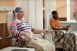 © Seventyfour - Portrait of young woman listening to music while getting chemotherapy treatment in clinic sitting in chair with IV drip copy space
