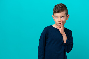 Wall Mural - Little kid boy isolated on blue background whispering something with surprise gesture while looking to the side