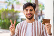 © luismolinero - Handsome Arab man holding a wallet at outdoors smiling and showing victory sign