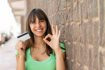 Young woman holding a credit card at outdoors showing ok sign with fingers