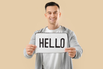 Canvas Print - Young man holding paper speech bubble with word HELLO on beige background