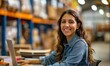 © LoLArt - Happy hispanic or latino female young woman warehouse worker sitting at desk with her laptop looking at camera and shelves with cardboard boxes in the logistic distribution delivery centre background