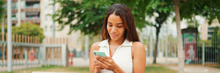 Wall Mural - Beautiful girl with long dark hair wearing white top sits on bench and uses mobile phone, Panorama. Young smiling woman writing in social networks on cellphone