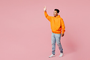 Wall Mural - Full body side profile view young man of African American ethnicity he wears yellow hoody casual clothes walk go waving hand isolated on plain pastel light pink background studio. Lifestyle concept.