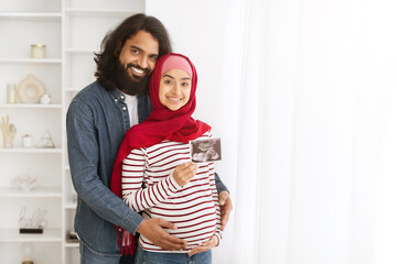 Wall Mural - Pregnant Muslim Couple Holding Baby Ultrasound Scan While Standing In Home Interior