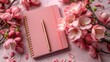 a pink notebook with a pen sitting on top of it next to a bunch of pink flowers on a table.