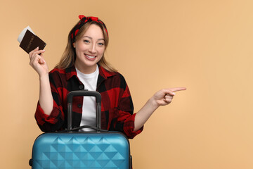 Wall Mural - Happy young woman with passport, ticket and suitcase pointing at something on beige background, space for text