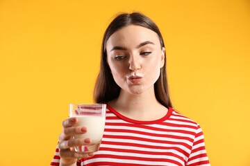 Wall Mural - Beautiful woman with milk mustache holding glass of tasty dairy drink on orange background