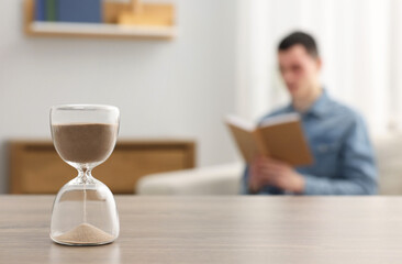 Wall Mural - Hourglass with flowing sand on desk. Man reading book in room, selective focus