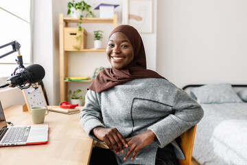 Portrait of young black woman in muslim headscarf smiling at camera sitting at desk in her room. Youth and ethnic people concept.