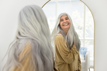 Cheerful carefree attractive senior Latin woman with long grey hair looking in mirror, smiling, laughing, enjoying reflection, wellness, dancing, posing with flying hair