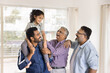© fizkes - Cheerful Indian daddy, granddad and great grandpa playing with little kid girl sitting on dads shoulders, laughing, standing close together at home, having fun, talking, enjoying family playtime