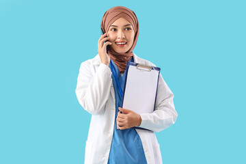 Sticker - Female Muslim doctor with clipboard talking by mobile phone on blue background