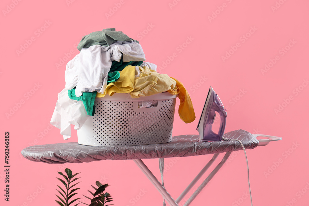 Electric iron with laundry basket on board against pink background