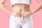 Fototapeta Panele - Young woman trying to button tight pants on pink background, closeup. Weight gain concept