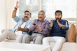 © fizkes - Excited happy adult Indian men of three family generations watching sport match on TV, celebrating team winning, laughing, shouting, waving hands, using remote control device, sitting on couch