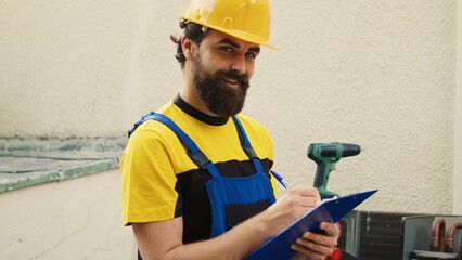 Wall Mural - Portrait of certified mechanic commissioned for outdoor air conditioner routine checkup, writing report on clipboard. Cheerful engineer doing hvac system inspection, looking for refrigerant leaks