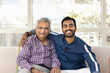 © fizkes - Positive adult Indian grandson embracing elderly grandad, giving family support, looking at camera with toothy smile, laughing, posing for home portrait, promoting elderly care