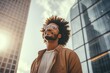 African American man with modern clothing, afro hairstyle, and sunglasses looks at the sky in the city center.