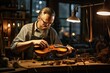 An adult craftsman wearing glasses sits at a table in a luthier outfit and paints a wooden violin in the light.