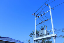 Electrical Transformer On Pole. High Voltage Transformer On Twin Concrete Poles With Cables And Fuse Blocks On Blue Sky Background In Bottom View With Copy Space With Selective Focus.