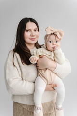 Wall Mural - Portrait of mommy embracing baby girl with bow isolated in background white wall. Mom holds in her hands 6 month old little girl. Cute baby in mom's arms closeup. Mother hugging his daughter.