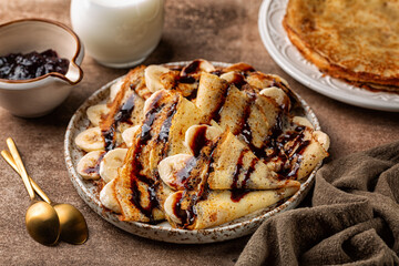 Poster - Homemade gluten-free crepes made with rice flour. Served with banana and chocolate sauce on a brown table surface. Milk and jam on background. Breakfast time.