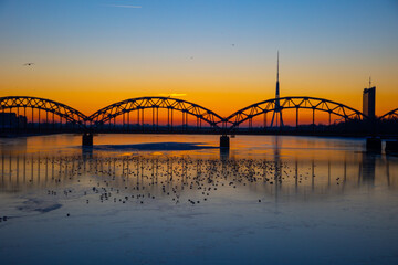 Wall Mural - A beautiful sunrise scenery with iron bridge over the frozen river Daugava in Latvian capital city Riga. Winter landscape of Northern Europe.