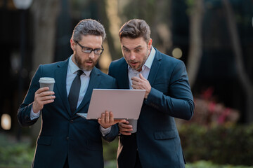 Wall Mural - Two surprised businessman using laptop outdoor. Handsome business man in suits working on laptop and communication with business projects in city. Partners thinking and plan for business.