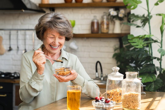 Cheerful senior grandmother eating cereals for breakfast mixing with berries drinking apple juice at home kitchen. Healthy dieting concept. Active seniors