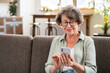 Cheerful caucasian senior woman using cellphone while relaxing on the sofa couch at home. Online shopping, scrolling social media, paying bills in mobile application